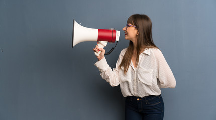 Woman with glasses over blue wall shouting through a megaphone to announce something in lateral position