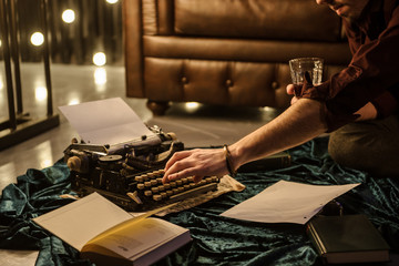 lateral view of man's hand typing on vintage typewriter on a piece of newspaper and dark blue velvet fabric on the floor in a dark room with light bulbs near a brown leather sofa