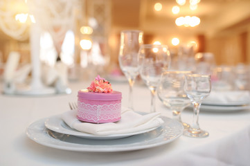 close up photo of knife and fork in a folded napkin on two plate near a set of glasses and a pink round gift box with a flower on the top on the napkin