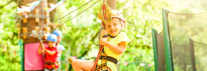 Brave young girl in helmet climbs on tree tops in amusement rope park on summer holidays, children...