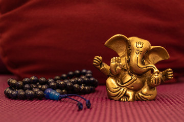 Golden statue of Ganesha with blue mala beads on yoga mat in front of a yoga pillow. Close up of Japa Meditation accessories.
