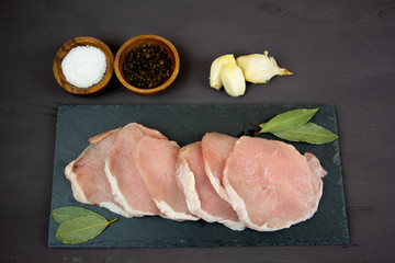 horizontal photo of whole pork tenderloin raw meat is on a black slate dish with pepper salt aromatic herbs and white garlic