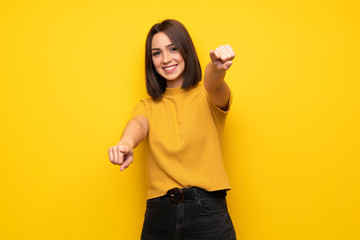 Young woman over yellow wall points finger at you while smiling