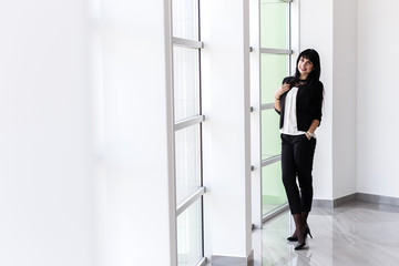Young  happy brunette woman dressed in a black business suit standing near the window in a office, smiling, looking at camera.
