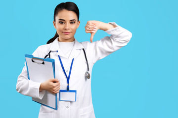 Portrait of bright young woman doctor with stethoscope and clipboard show bad gesture with hand over blue background.