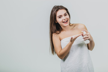 Fototapeta premium Model look is a beautiful young twin in a white terry towel. Applying a body lotion on her shoulder and smiling while standing on a white background.