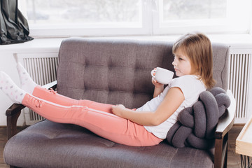 Cheerful cute adorable girl drinking milk in a cup at home in a sofa