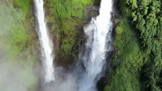 Upward drone shot of the Tad Fane waterfall in Laos surrounded by jungle, with steam going up from the bottom of the waterfall, on a sunny day with some clouds