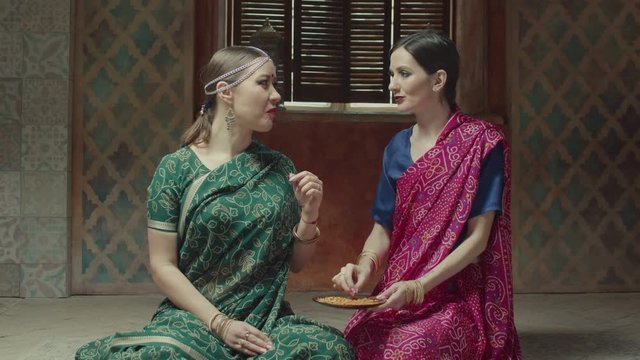 Seated on floor attractive women in hindu dress sari with bindi and traditional jewels eating with hands. Lovers of indian cuisine enjoying flavor of ethnic delicious meal, Bombay mix with appetite.