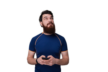 Photo of bearded man dressed in blue tshirt, holding mobile while thinking and looking  up against white background