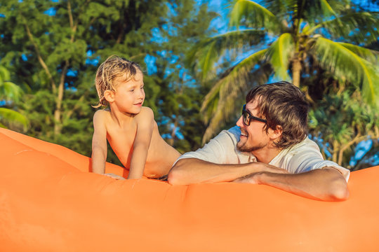 Summer lifestyle portrait of father and son sitting on the orange inflatable sofa on the beach of tropical island. Relaxing and enjoying life on air bed