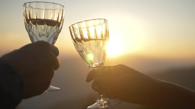 Couple holding glass of wine, making a toast over sunset. Slow motion shot of cheers with wine glasses, mountain setting.