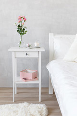 Bedroom interior with white furniture and grey concrete wall. Light female modern stylish bedroom. Bedside table, vase with rose flowers, lighted candle, pink box, bed with white linen, fluffy carpet.