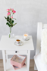 Bedside table, vase with rose flowers, lighted candle and cup of tea. Light female modern stylish and cozy bedroom. Bedroom interior.