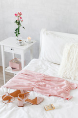 Elegant long pink dress, heeled sandals and perfume lying on the bed. Girl choosing outfit. Female modern stylish and cozy bedroom. Bed with white linen, bedside table, vase with flowers, candle.