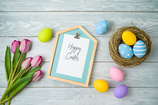Easter holiday background with easter eggs in bird nest, note and tulip flowers on wooden table.