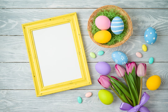 Easter holiday background with easter eggs in basket, photo frame and tulip flowers on wooden table.