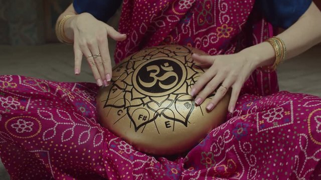 Midsection of seated in lotus pose woman playing traditional indian music using glucophone decorated with mantra symbols. Dressed in sari female drumming rhythm on hank drum by hands with bracelets.