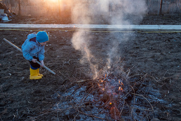 Two happy children in blue work clothes and their young slender mother burn old grass in garden. Family is happy together. Bonfire in front of sunset on a warm spring evening.