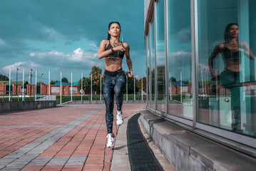 Beautiful young athletic girl tattoos runs morning for jog, fitness training, sportswear, leggings top. Phone listens music headphones. Summer city workout. Background sky glass windows. High jump.