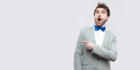 Portrait of amazed handsome bearded man in casual grey suit and blue bow tie standing looking at camera with surprised face and pointing at copyspace. studio shot, isolated on light grey background.