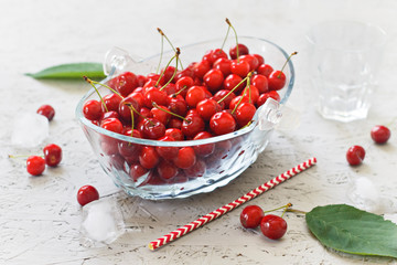 Cherry berries with leaves in a glass bowl and ice cubes on a gray background. Side view