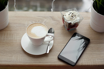 White cup of hot cappuccino on white saucer, black mobile phone, red velvet dessert and green grass in pots on wooden bar table next to window