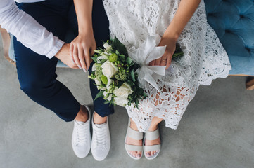 Bride and groom with a wedding bouquet of white roses are sitting side by side on the couch and holding hands. Lace dress, studio, white shoes and sandals.
