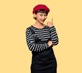 Young woman with beret making phone gesture. Call me back sign over yellow background