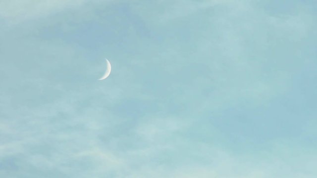 Slim crescent moon hangs in a blue sky with wispy clouds in daytime