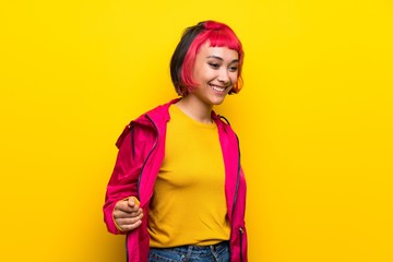 Young woman with pink hair over yellow wall posing with arms at hip and smiling