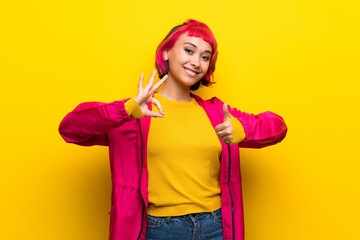 Young woman with pink hair over yellow wall showing ok sign and thumb up gesture