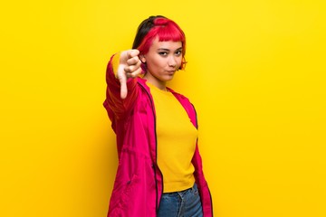 Young woman with pink hair over yellow wall showing thumb down with negative expression