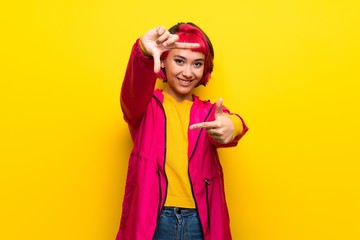 Young woman with pink hair over yellow wall focusing face. Framing symbol