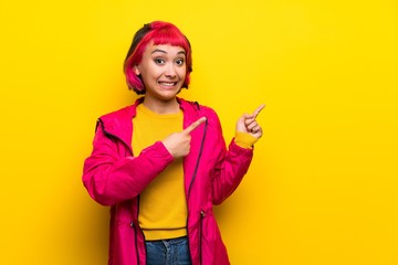 Young woman with pink hair over yellow wall frightened and pointing to the side