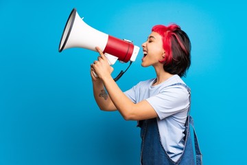 Young woman with pink hair over blue wall shouting through a megaphone