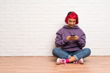 Young woman with pink hair sitting on the floor sending a message with the mobile