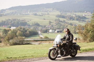 Long-haired bearded cool biker in sunglasses and black leather clothing riding modern cruiser motorcycle along sunny asphalt road on bright summer day on background of green rural misty landscape.