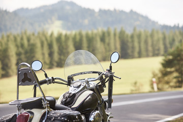 Detail of black shiny powerful cruiser motorcycle parked on roadside on blurred sunny outdoors background. Modern transportation technology, speed and long-distance traveling concept.