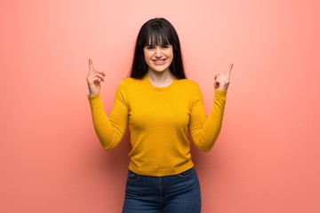 Woman with yellow sweater over pink wall making rock gesture