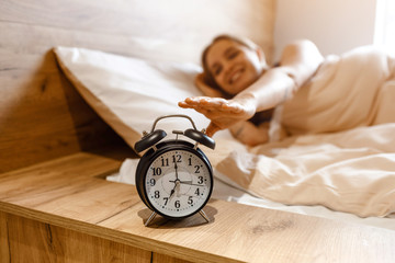 Young beautiful blonde woman lying in bed in morning. Positive happy model reaching alarm clock...