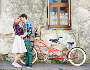 Young attractive romantic tourist couple, bearded man and blond woman standing together at modern tandem bicycle on lit by bright sun empty pavement at cracked wall of old residential house.