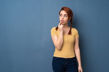Young redhead woman over blue background having doubts while looking up