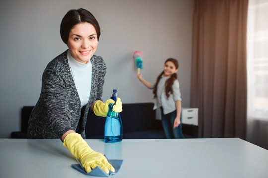 Beautiful Brunette Caucasian Mother And Daughter Cleaning Together In Room. Cheerful Young Woman Washing Desk And Hold Spray. Girl Stand Behind And Blust The Dust.