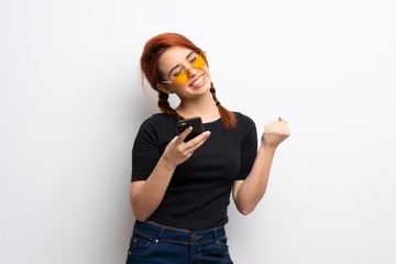 Young redhead woman over white wall with phone in victory position