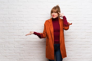 Woman with coat over white brick wall making the gesture of madness putting finger on the head