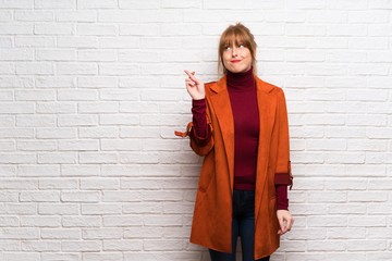 Woman with coat over white brick wall with fingers crossing and wishing the best