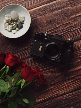 Red roses lying next to vintage camera and pieces of jewelry on wooden desk background. Flat lay. Top view