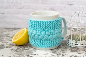 White mug on the table is dressed in a knitted frame. Yellow lemon