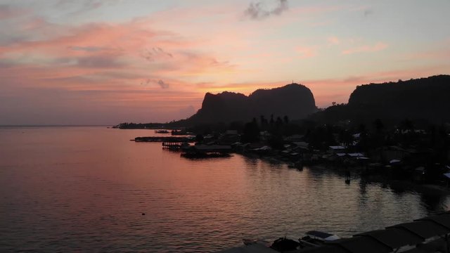 Sunset colors fill the sky and reflect on the calm surface of the sea. A marina on the coast of Tawi Tawi, Philippines filled with boats for the night and an empty pier. Homes along the shore.